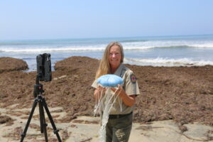 An interpretive ranger stands on the beach smiling at the camera, holding a model of a blue sea jellie, with a videoconferencing device on a tripod to her left. The waves in the background are crashing on the shore.