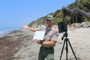 An interpretive ranger stands on the beach with the water on the left and the bluffs on the right. He is smiling and motioning his hand toward the beach while holding a whiteboard with a drawing of a watershed and a smiling sun. Next to him is his video conferencing device on a tripod.