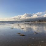 Image shows the ocean on the left with a wet sand covering the beach, reflecting the sky and clouds in the distance. Some clumps of kelp sit on the wet sand. Mountains are nestled under the clouds in the distance.