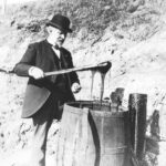 Black and white photo of a gentleman using a long stick to pull tar from a barrel.
