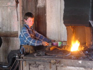 a boy in a blue and grey flannel shirt pokes a fire at the blacksmith shop