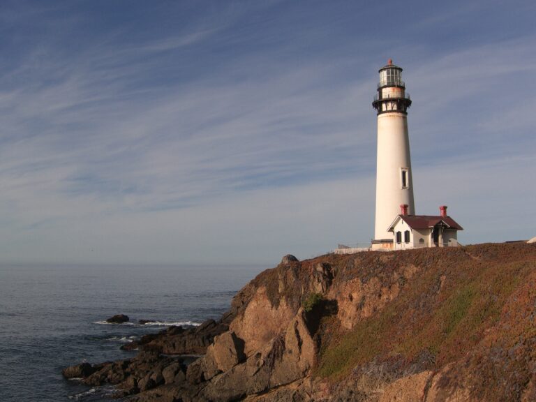 a white lighthouse with small white house with red roof and two chimneys on the edge of a cliff with the ocean on the left.